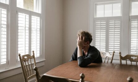 A man sits alone at a table in a bright room, displaying deep contemplation.