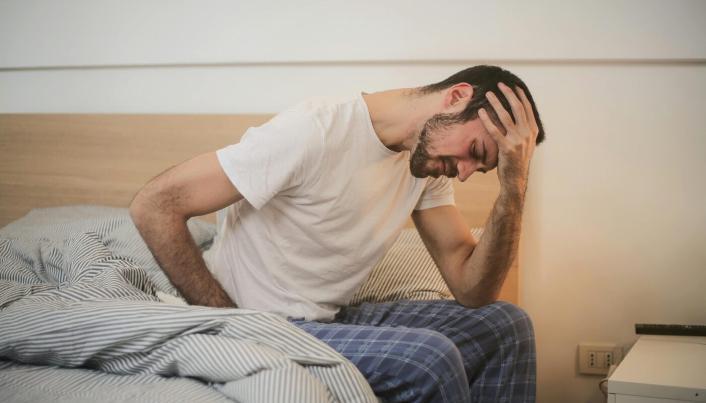 A young man in pajamas holding his head, sitting on a bed, appears to be experiencing a headache.