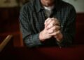 A man in a checked shirt praying in a church, hands clasped together.